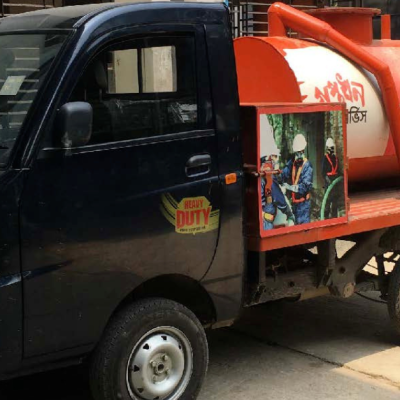 A fecal sludge management vacuum truck parked on the side of a paved road in Bangladesh, with two workers standing beside it. The truck is fitted with large hoses and equipment used for emptying septic tanks and pit latrines. Surrounding the vehicle are small buildings, trees, and utility poles, reflecting a typical urban neighborhood setting. The image illustrates formalized FSM service operations in practice.