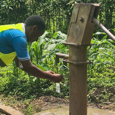 A field enumerator wearing a reflective safety vest collects a water sample from a hand pump in a rural setting in Kabarole District, Uganda. The water sample is part of the Heavy Metals Water in Kabarole study, which examines the presence of chemicals and heavy metals in drinking water sources. The background features lush green vegetation and maize plants, while another individual operates the pump handle to release water into the sample bottle.