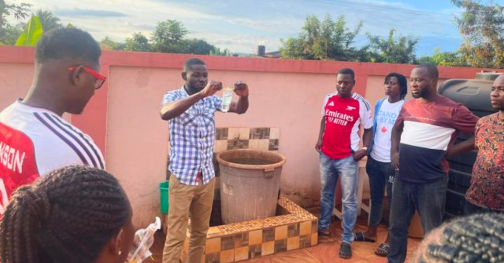 A group of enumerators in Asutifi North conducting water quality tests on drinking water. One individual in a checkered shirt is demonstrating the test procedure while others observe attentively. The setting includes a water storage container and a black tank in the background. Heavy Metals Water Asutifi North.