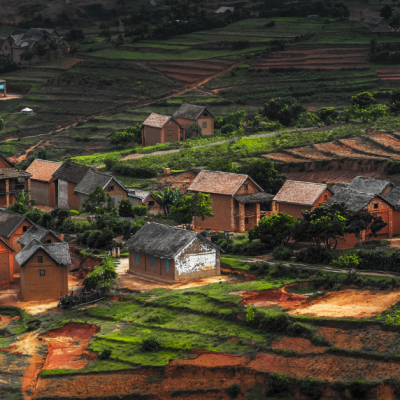 A small village on the hill with green gardens and red soil in Madagascar: This image showcases a rural village situated on a hilly landscape with green fields, tiered farmlands, and patches of red soil. The village consists of several small houses built with reddish-brown materials, clustered together among lush greenery. The surrounding fields display agricultural activity, with footpaths weaving through the landscape. The distant hills, dotted with additional houses, complete the scenic rural view. Water supply in Madagascar