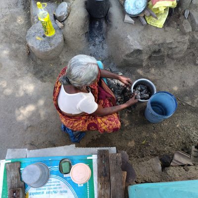 An elderly woman in a red and orange sari prepares food over a traditional outdoor stove in Tamil Nadu, India. She is seated on the ground, tending to a pot of fish near a blue water bucket.